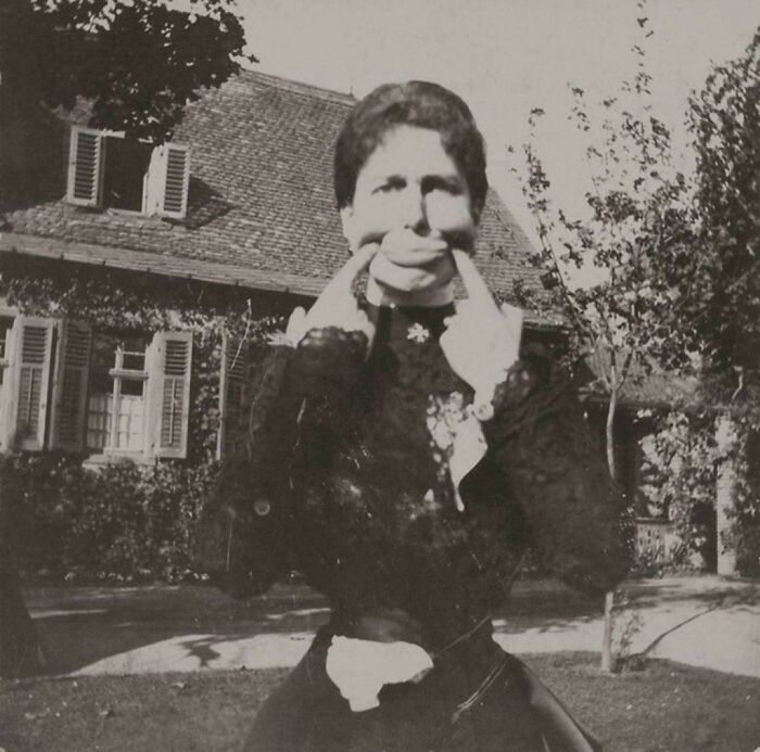 Victorian era woman making a funny face by pulling her mouth in a garden with a vintage house in the background.