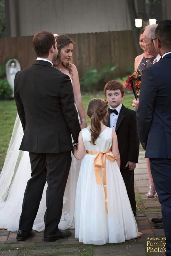 Young boy in tuxedo making an awkward face during wedding ceremony with bride, groom, and children present outdoors.