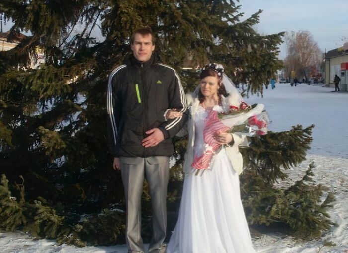 Bride in white wedding dress holding flowers with groom in casual jacket posing outside near a tree awkward wedding photo