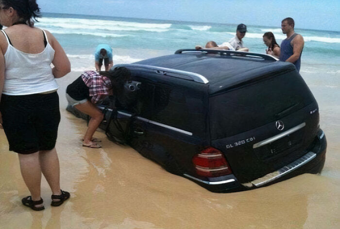 Black luxury car partially submerged in ocean sand with people inspecting it after an expensive fail.