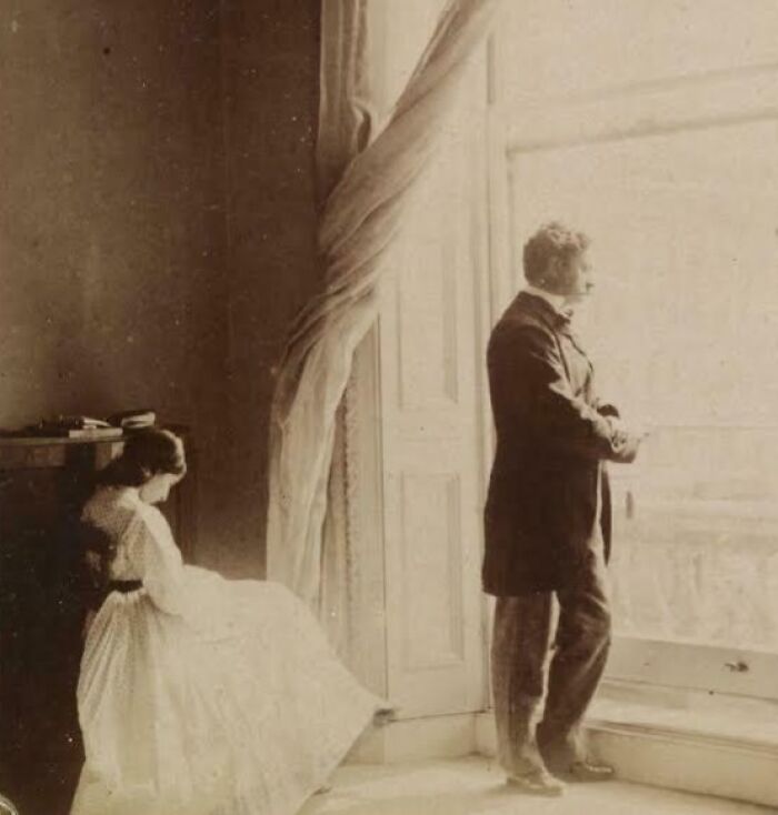 Victorian era couple in period clothing by a large window, showcasing lifestyle and fashion from the Victorian era.