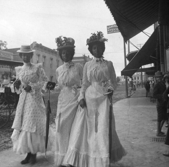 Three Victorian era women wearing elegant dresses and hats walking on a town sidewalk holding umbrellas and accessories.