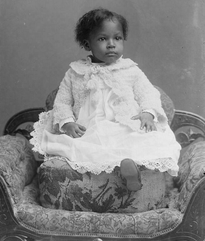 Young Victorian era child dressed in lace and sitting on a patterned Victorian armchair from the Victorian era.