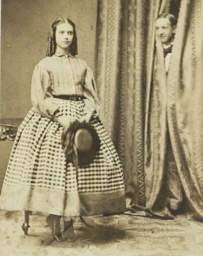 Victorian era woman in patterned dress holding a hat while a man peeks from behind heavy curtains in vintage photo.