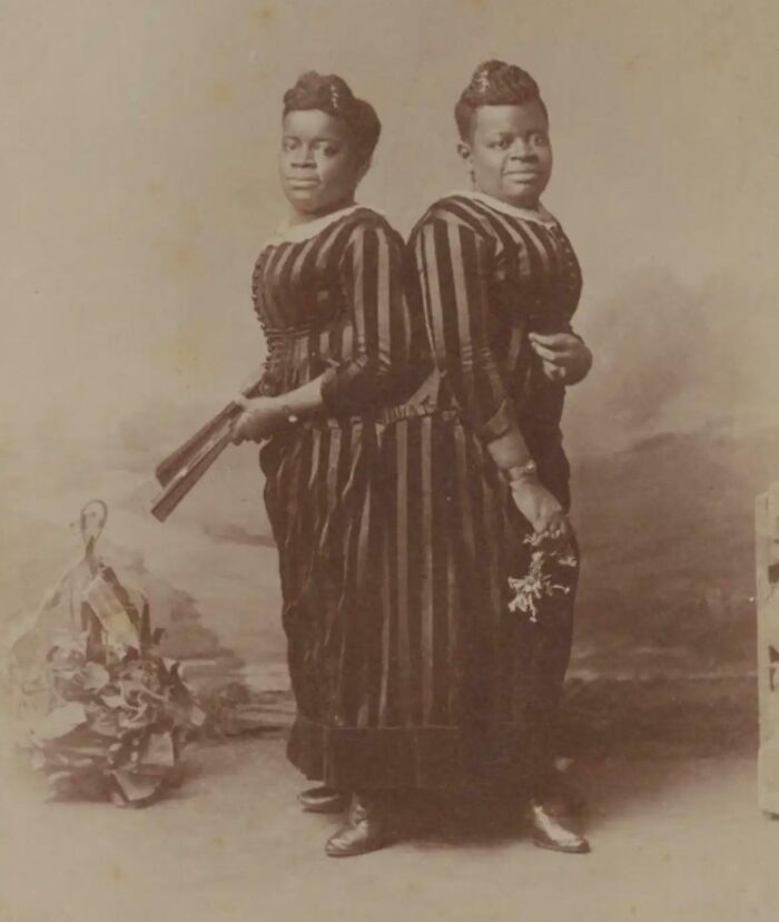 Two Victorian era women in striped dresses posing back-to-back, one holding a fan, the other holding flowers in a studio setting.