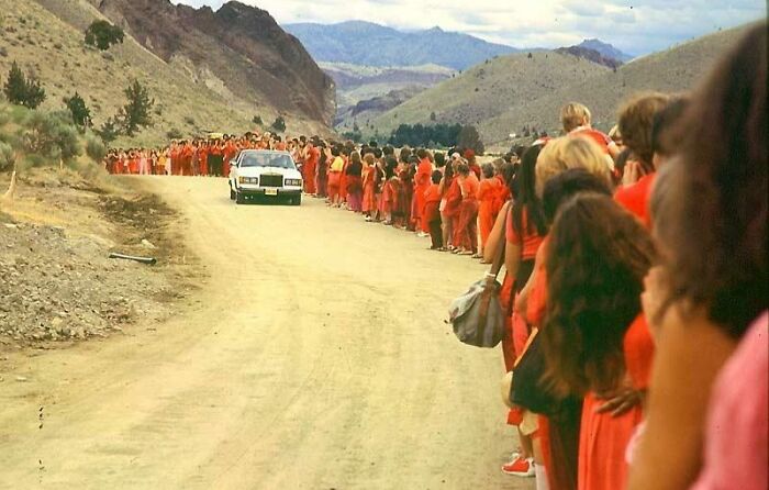 Crowd of women in red dresses lined up along a dusty road with a car approaching, evoking unsettling history and crime.