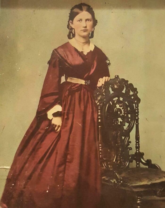 Victorian era woman in a maroon dress standing next to an ornate wooden chair in a vintage photograph.
