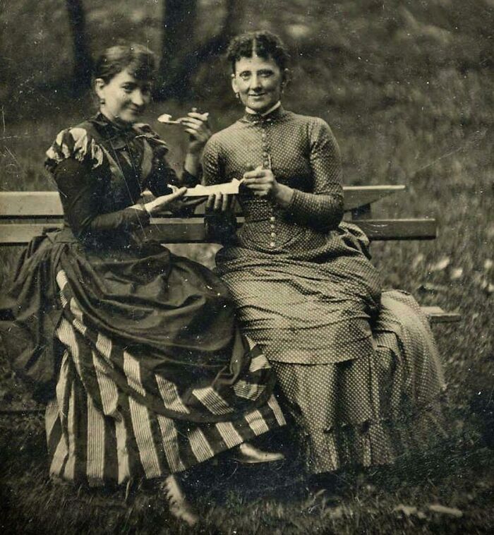 Two Victorian-era women in period dresses sitting on a bench outdoors, sharing a meal in a candid moment.