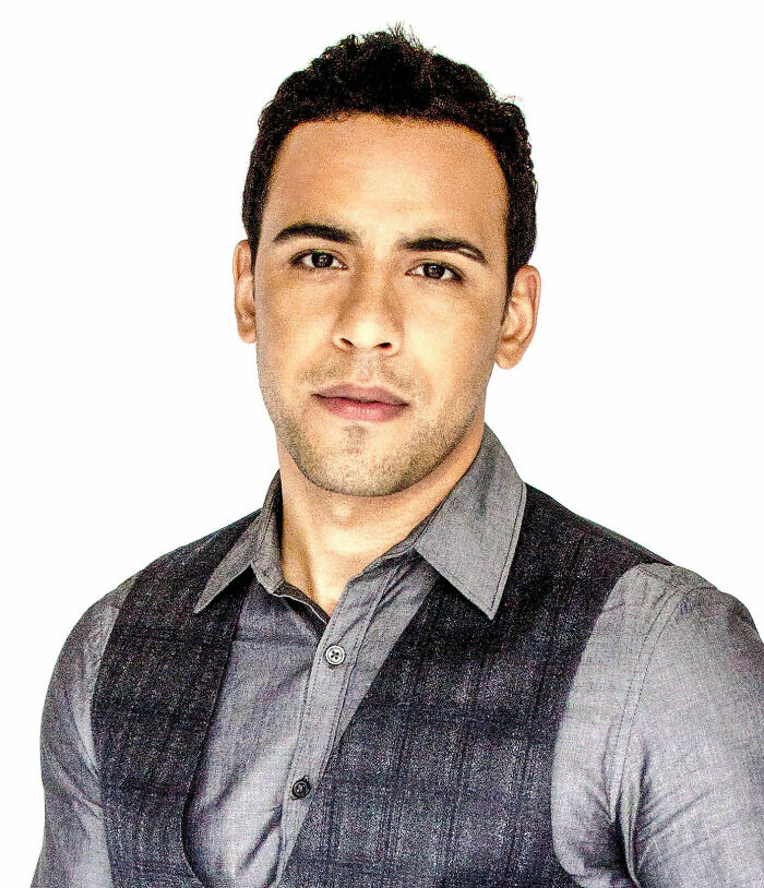 Victor Rasuk in a gray shirt and dark vest, posing against a plain white background for a professional portrait.