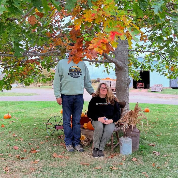 Tall person standing next to seated woman under tree with autumn leaves, pumpkins on grass highlighting height struggles.