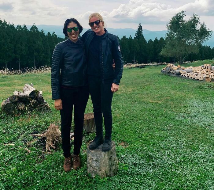 Two tall people outdoors, one standing on a tree stump to match height, wearing black jackets and sunglasses.