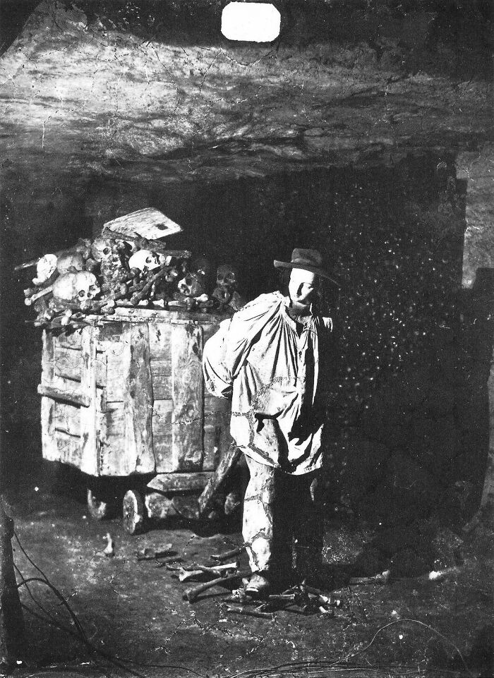 Historic site photo showing a figure standing next to a cart filled with human skulls and bones in a dim underground setting.