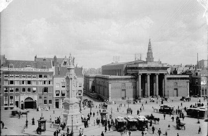 Historic site photo showing a busy square with vintage buildings, a monument, and early 20th-century vehicles and pedestrians.