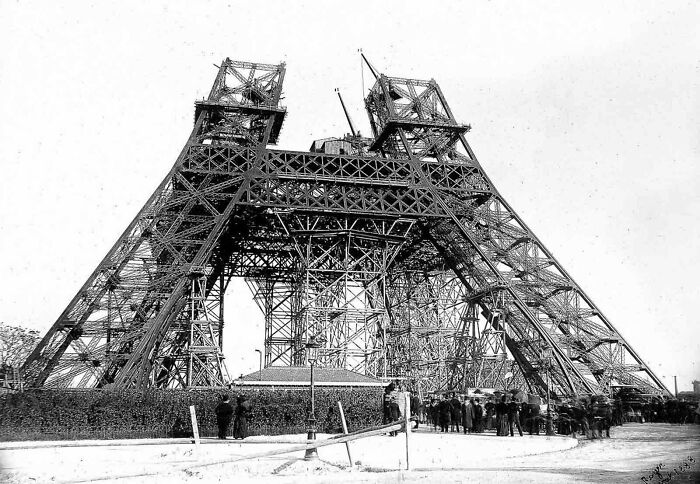 Historic site photo showing early construction phase of the Eiffel Tower with scaffolding and workers gathered around.