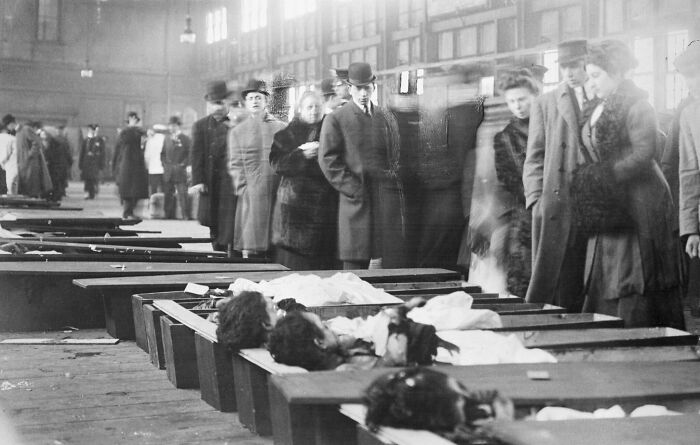 Black and white historic site photo showing people in early 20th century attire viewing open coffins in a large hall.