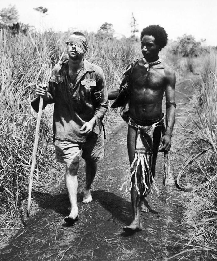 Historic site photo showing two men walking barefoot on a dirt path surrounded by tall grass in a rural setting.
