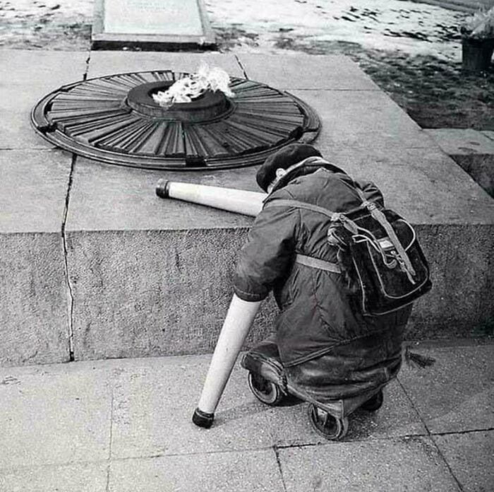 Child kneeling by an eternal flame at a historic site, wearing a backpack and holding rolled-up paper.