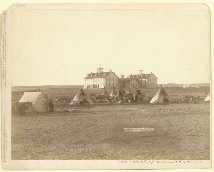 Historic site photo showing a 19th-century U.S. school for Indians with tents and carts in the foreground.