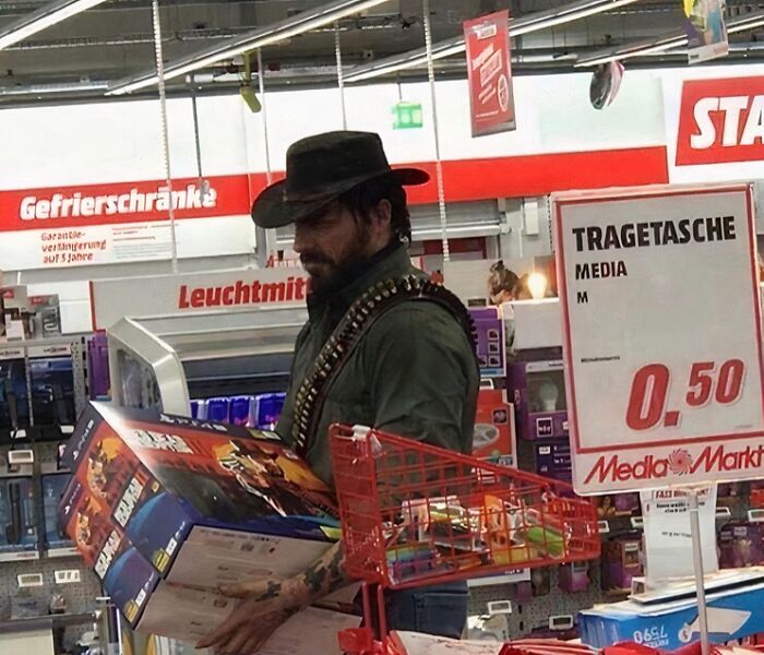 Man dressed as cowboy holding boxed video games inside electronics store, a confusing picture that is funny to see.