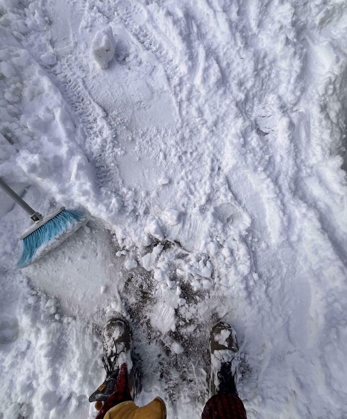 Person standing in snow with boots covered in snow next to a broom and partially shoveled path showing unfortunate fail.