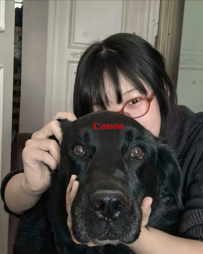 Woman hiding behind a black dog with a Canon logo on its forehead in a confusing picture that is funny for no clear reason