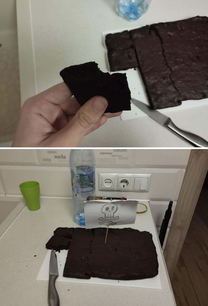 Hand holding a burnt brownie piece with a blackened dessert and a pirate flag on a kitchen counter showing ruined dessert.