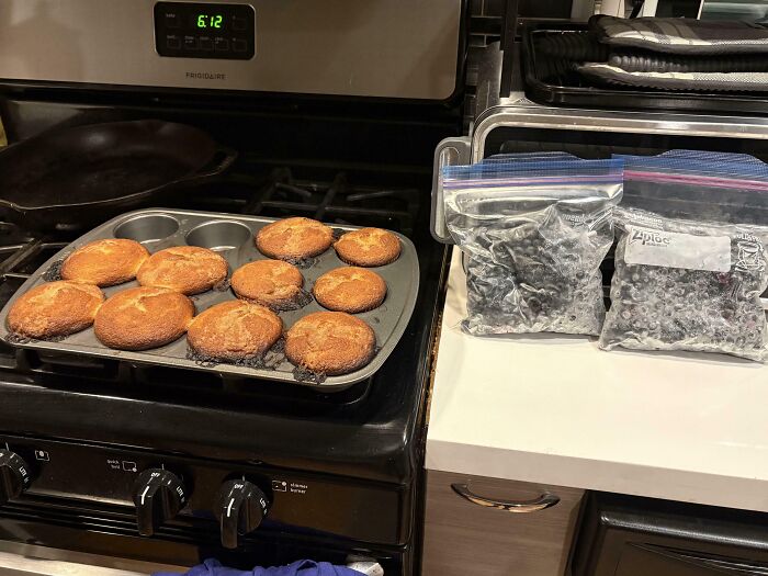 Overbaked muffins with burnt edges cooling on stove next to bags of blueberries, showing dessert ruined unexpectedly.