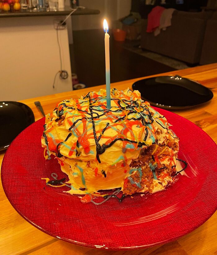 Cake ruined with messy frosting and colorful drizzles, topped with a single lit candle on a red plate.