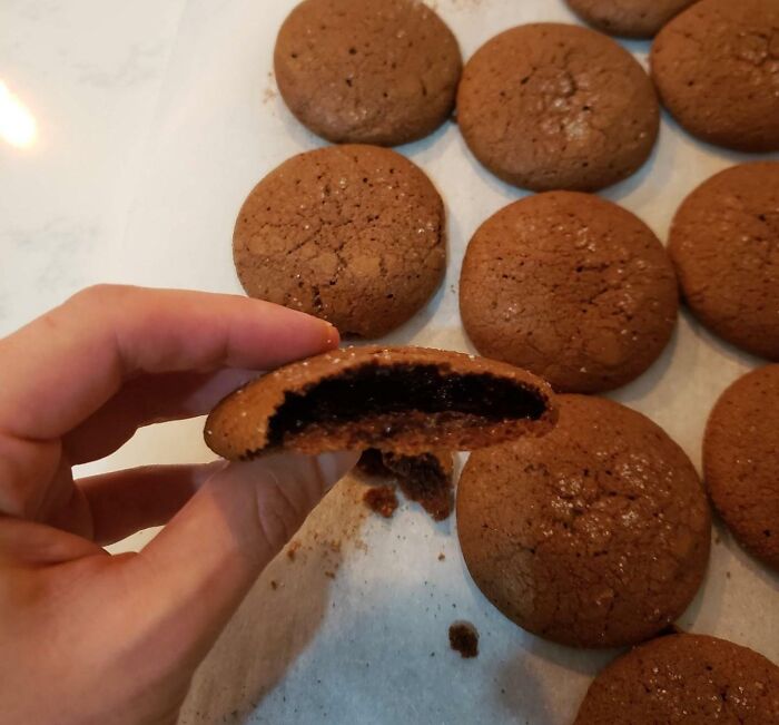 Hand holding a chocolate cookie broken in half showing undercooked gooey center among more cookies on a tray, dessert ruined.