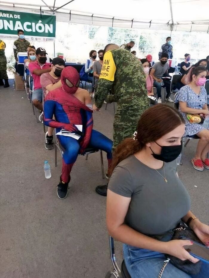 Person dressed as Spider-Man receiving vaccine shot while others wait in line, a confusing and funny picture at a vaccination site