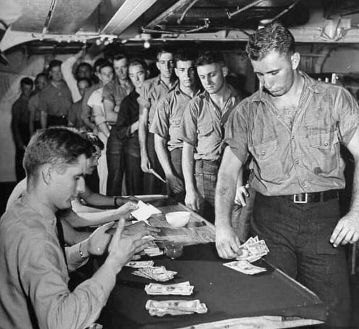 US history photo of men in military uniforms lining up to receive money during World War II on a ship’s lower deck.