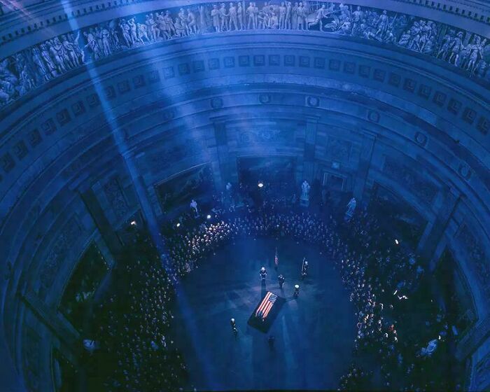Interior view of a historic US building with a flag-draped coffin and crowds paying respects in a solemn ceremony.