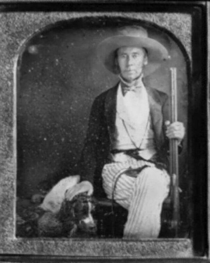 Vintage US history photo of a man in period clothing holding a rifle, seated with a dog beside him.