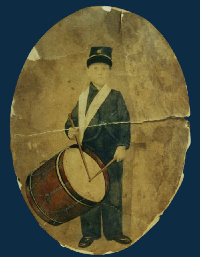 Vintage US history photo of a young drummer boy in uniform holding drumsticks and a large drum.