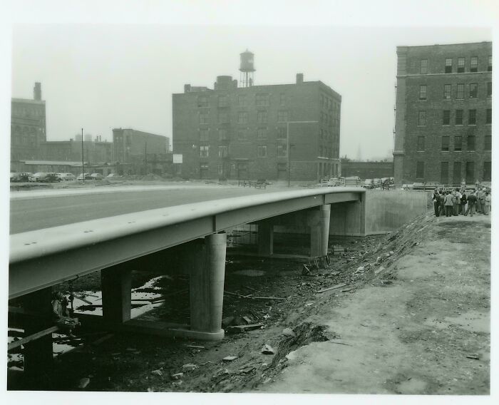 Black and white US history photo showing an urban bridge under construction with buildings and people nearby.
