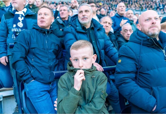 Young boy in green jacket among men at crowded stadium, street photos by Perry Hall capturing life exactly as it happens.