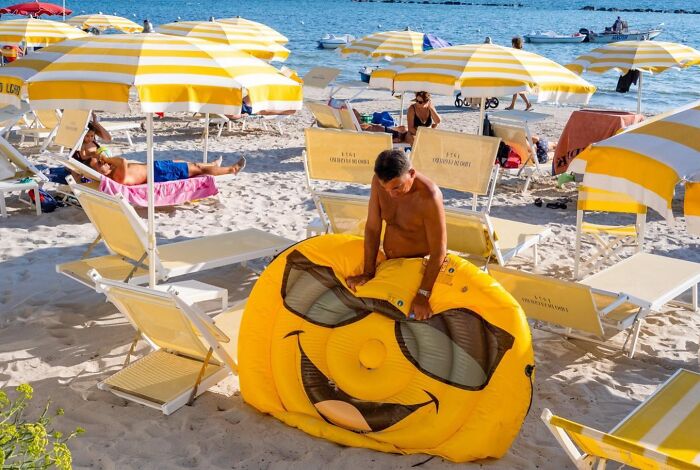 Man inflating a large smiley face floatie on a sunny beach with yellow and white umbrellas, street photos by Perry Hall.