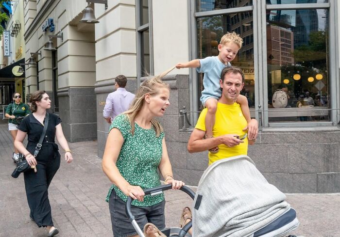 Family walking on city sidewalk with stroller and child on father's shoulders in vibrant street photo by Perry Hall