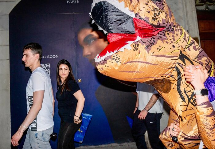 Young adults walking on city street, one wearing a large inflatable dinosaur costume in candid street photos by Perry Hall.