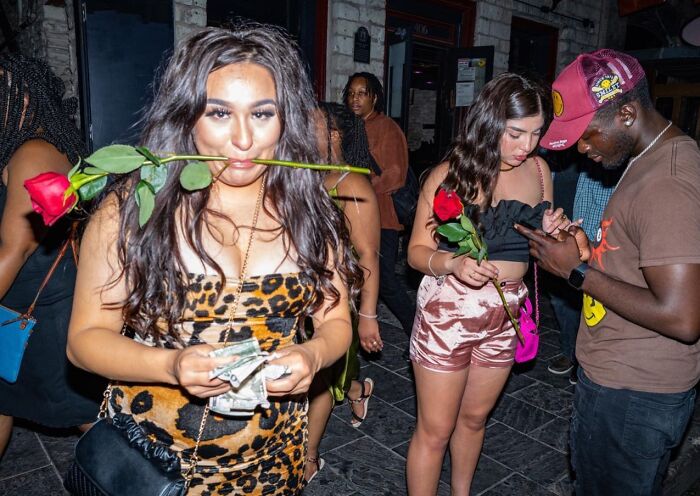 Young woman holding money with rose in mouth, surrounded by people, captured in candid street photos by Perry Hall.