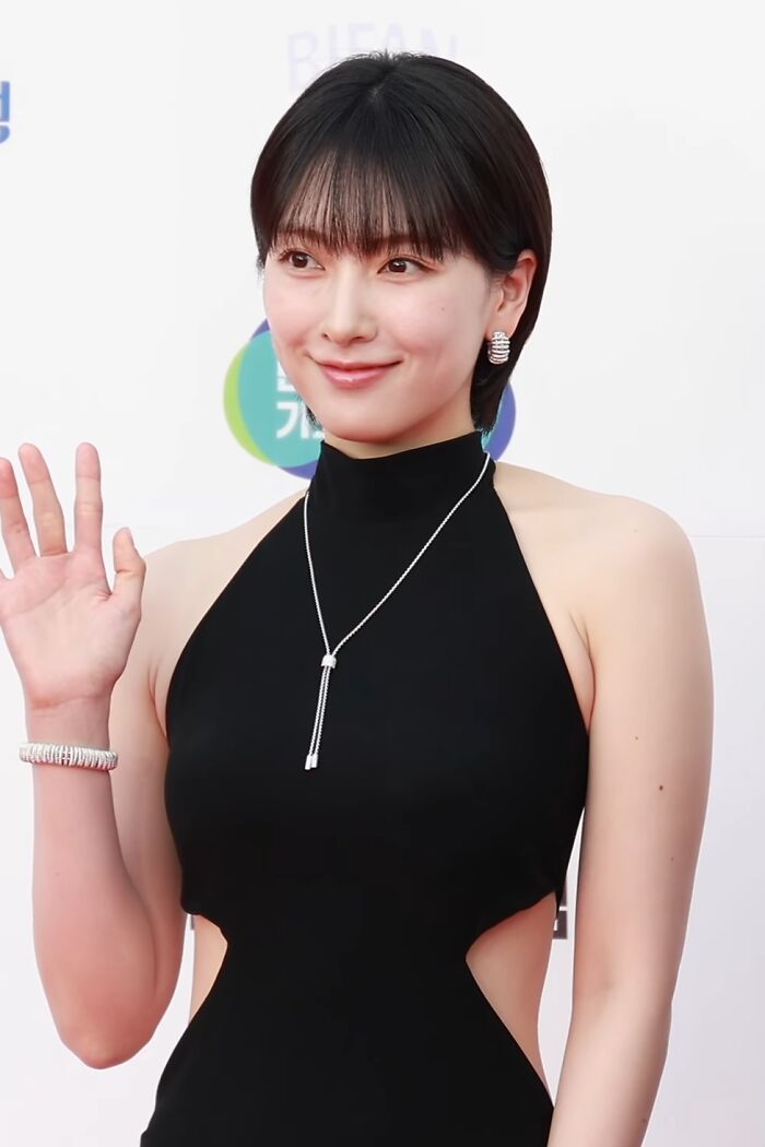 Kang Ji-young smiling and waving, wearing a black dress with cutouts and silver jewelry at a formal event.