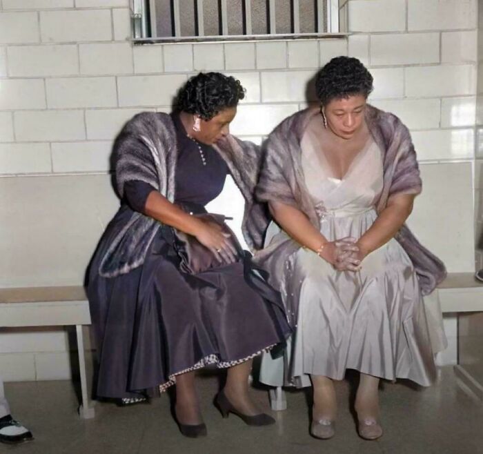 Two women in vintage dresses and fur stoles sitting on a bench in a historical photograph from the past.