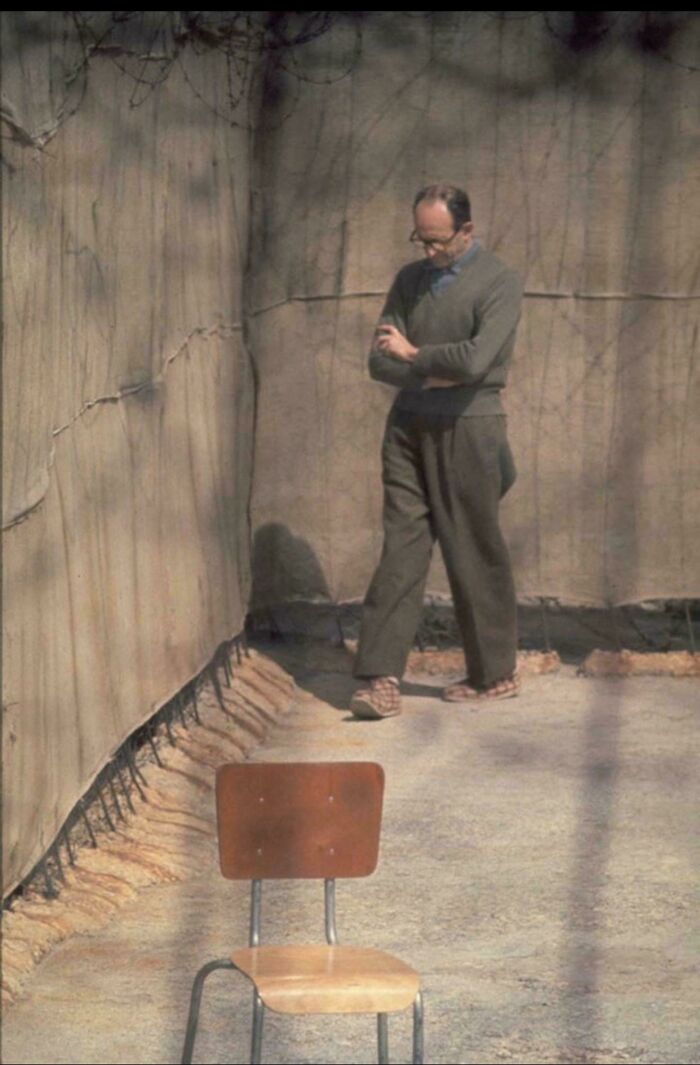 Man in vintage clothing walking beside a bare wall and empty chair in an interesting historical photograph.