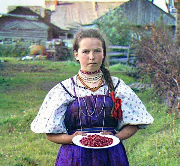Young girl in traditional dress holding a plate of berries in a rural setting, an interesting historical photograph.