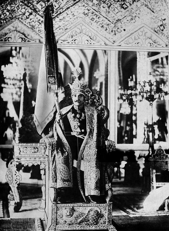 Black and white historical photograph of a regal figure seated on an ornate throne holding a ceremonial flag in a grand room.