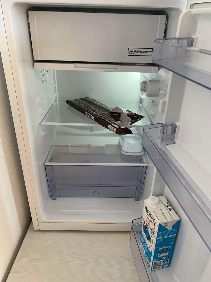 Open fridge with minimal contents including a partially eaten chocolate bar and a carton, showing unusual fridge contents.