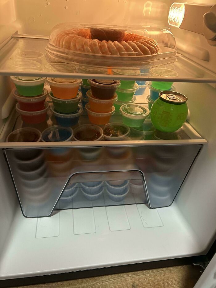 Fridge contents showing plastic-wrapped shrimp tray and colorful gelatin cups inside an illuminated refrigerator.