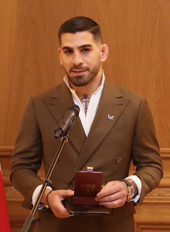 Ilia Topuria in a brown suit holding an award and speaking into a microphone in a formal indoor setting