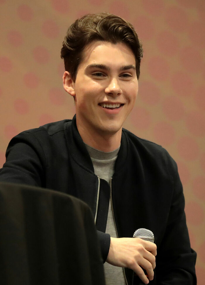 Jeremy Shada smiling and holding a microphone while speaking at an indoor event in a casual black jacket.