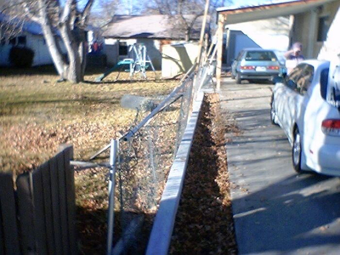 Poorly constructed DIY fence collapse next to driveway with fallen fence panels and scattered debris in a residential yard.